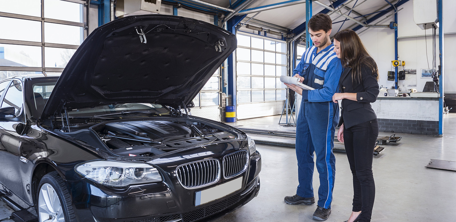 Mechanic with clipboard and and female customer standing to right of black BMW with bonnet open.